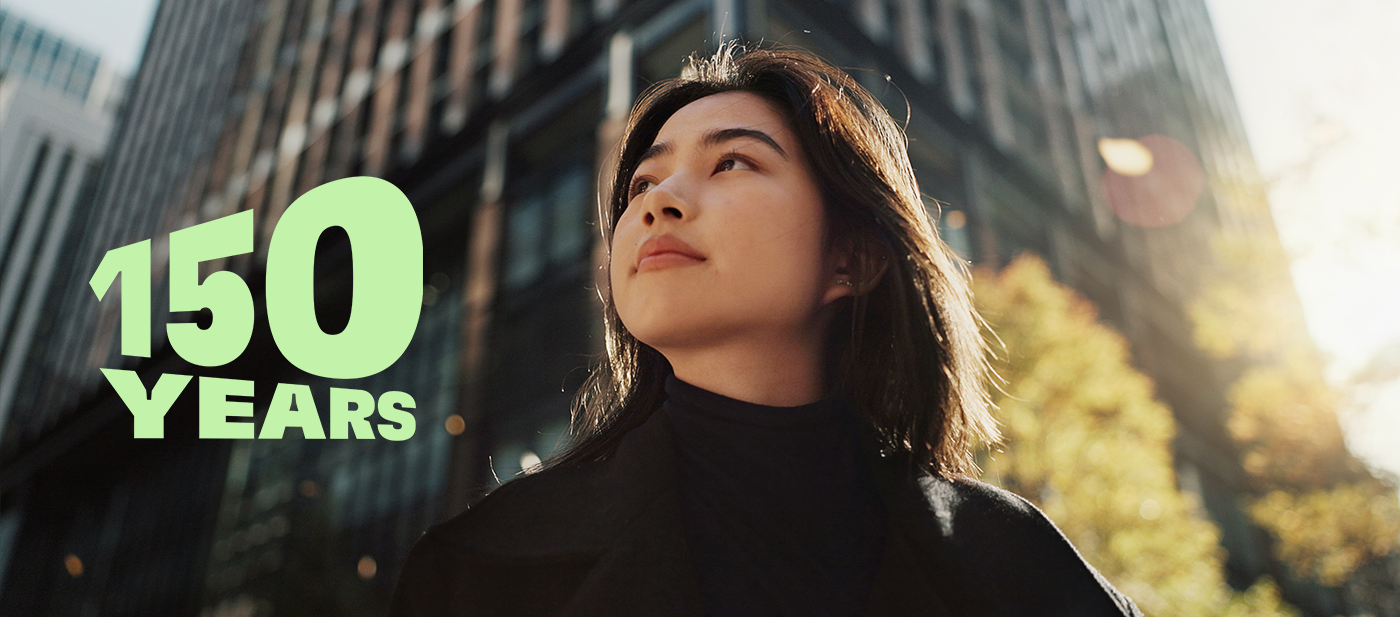  Grüner Text mit der Aufschrift „150 JAHRE“ über einer modernen Stadtlandschaft mit hohen Glasgebäuden und Herbstbäumen, die das 150-jährige Jubiläum symbolisieren.