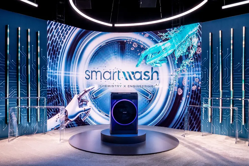 Eine futuristische Szene zeigt eine Waschmaschine auf einer kleinen, runden Bühne im Zentrum. Dahinter ist ein großer Bildschirm zu sehen, der eine Nahaufnahme einer Waschtrommel zeigt, sowie das Wort „smartwash" mit den Worten „CHEMISTRY x ENGINEERINGAI" darunter.