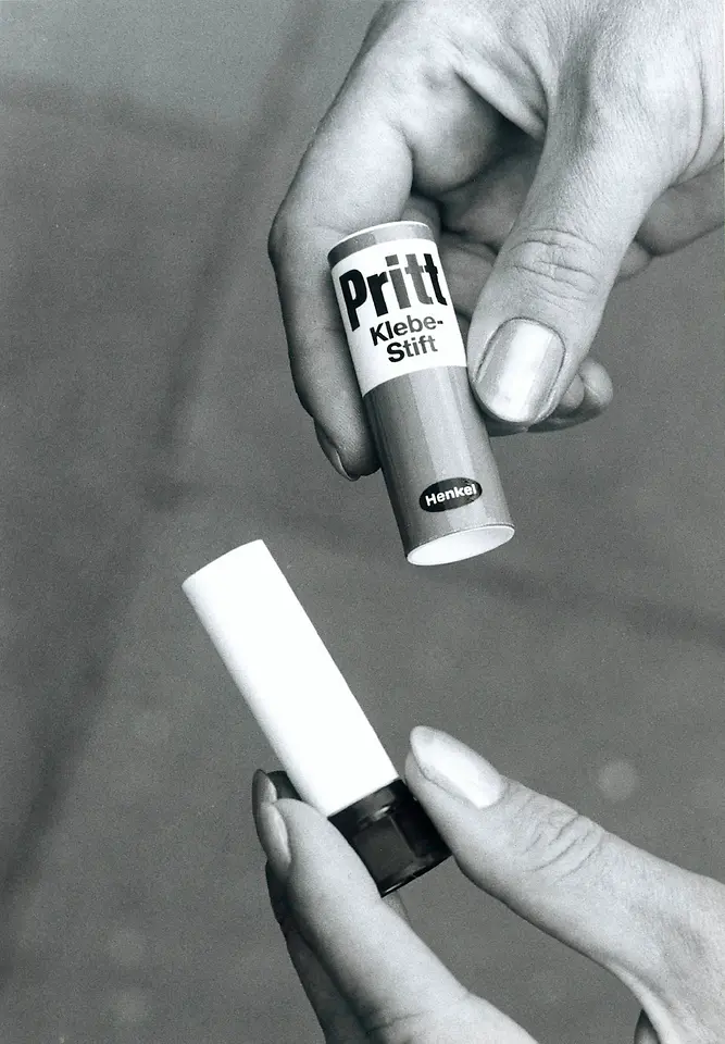 Schwarz-weiß-Foto: Zwei Hände halten einen Pritt-Klebestift. Eine Hand setzt die helle Kappe ab, die andere hält den Stift mit der Aufschrift „Pritt Klebe-Stift Henkel“