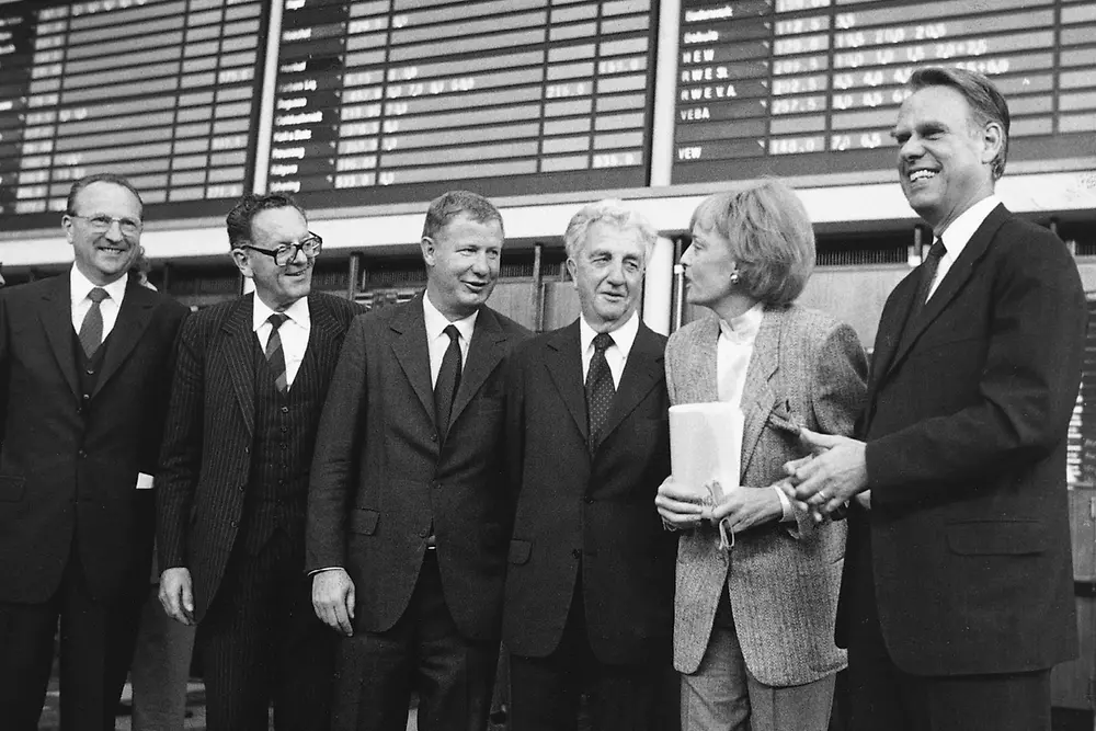 Schwarz-weiß-Fotografie von sechs Personen in Anzügen und einer Frau im Hosenanzug vor der Anzeigetafel der Börse. Die Gruppe lächelt in die Kamera.