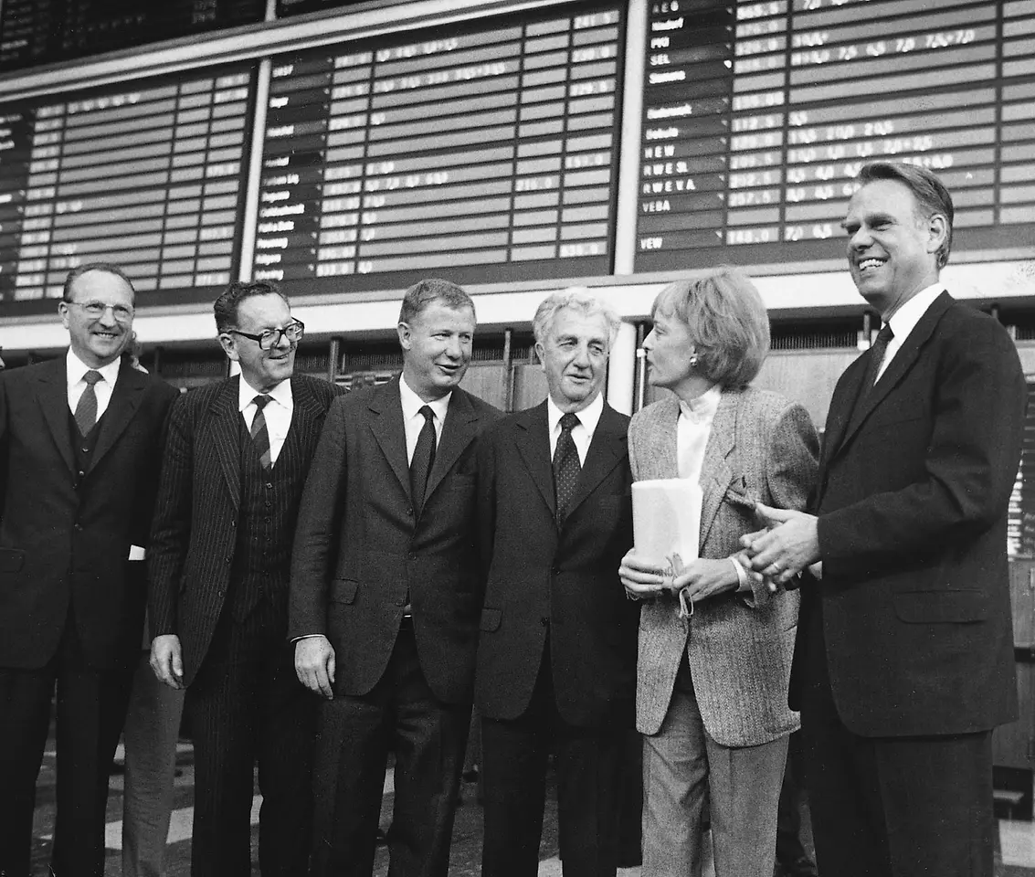 Schwarz-weiß-Fotografie von sechs Personen in Anzügen und einer Frau im Hosenanzug vor der Anzeigetafel der Börse. Die Gruppe lächelt in die Kamera.