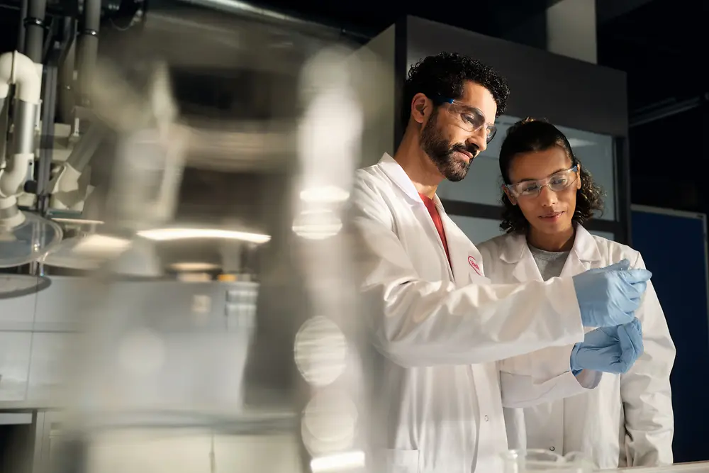 Zwei Wissenschaftler in weißen Laborkitteln, Schutzbrillen und Handschuhen untersuchen gemeinsam in einem Labor einen kleinen Probenbehälter.