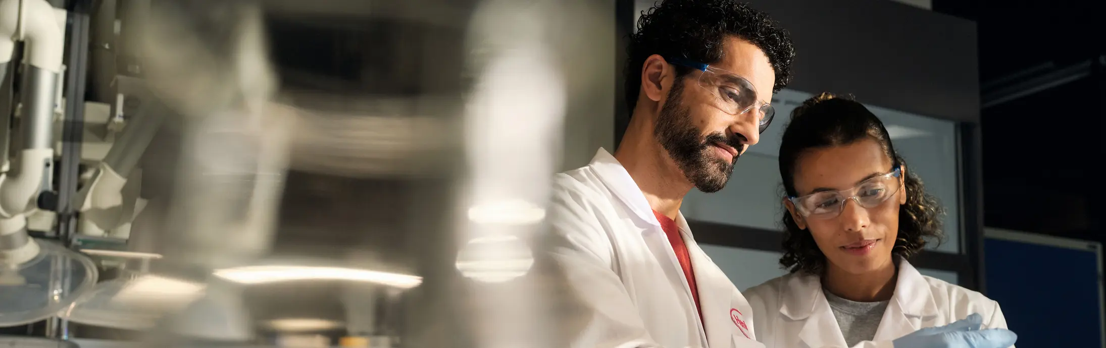 Zwei Wissenschaftler in weißen Laborkitteln, Schutzbrillen und Handschuhen untersuchen gemeinsam in einem Labor einen kleinen Probenbehälter.